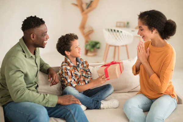 Family Holiday And Gift. Black Dad And Son Holding Birthday Present, Greeting And Surprising Mom Together, Sitting On Sofa At Home. Woman Receiving Wrapped Box From Husband And Kid