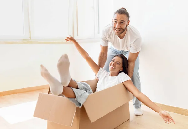 Relocation day, movement, real estate, mortgage. Happy handsome young man pushing cardboard box with girlfriend or wife cheerful woman inside, couple have fun while unpacking stuff in new house