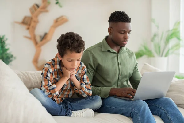 黑人年轻父亲忽略了他的不幸的小儿子使用笔记本电脑在网上工作坐在沙发上在家里 爸爸和可悲的小男孩有关系问题的概念 选择性焦点 — 图库照片