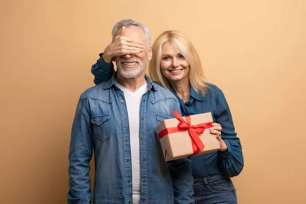 Beautiful cheerful blonde loving elderly woman making birthday surprise for husband, senior lady with gift box in her hand cover smiling man eyes, celebrating anniversary together, beige background