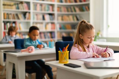 İlkokulun içerisindeki sınıfların masalarında oturan, not defterlerinde yazan ya da çizen çeşitli okul çocukları. Okula geri dön.