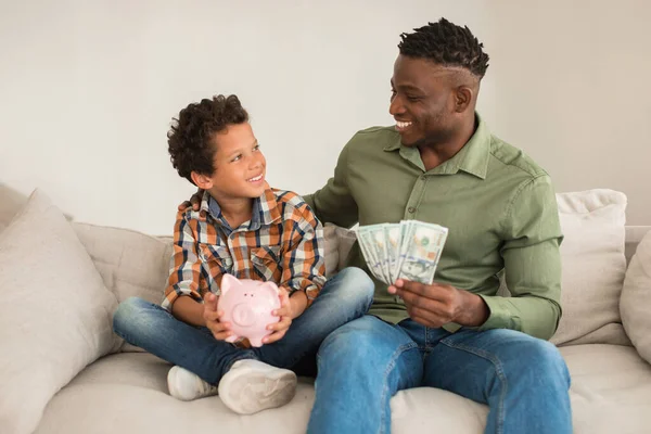 Personal Savings. Cheerful African American Dad And Little Son Holding Piggyback And Dollar Money Cash Indoor, Sitting On Couch And Smiling To Each Other. Financial Investments and Bank Offer Ad