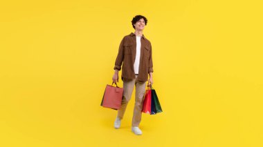 Satış, tüketim. Alışveriş yaptıktan sonra gülümseyen adam sarı arka planda poz veren renkli paketler, stüdyo çekimi. Müşteri alım teklifiyle ayakta duruyor. Panorama, tam boy