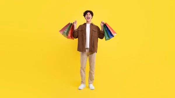 Yes, seasonal sales offer. Excited buyer guy raising hands with shopping bags, holding colored paper packages and expressing joy, shouting on yellow studio background, full length, panorama