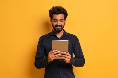 Sarı Stüdyo Arkaplanı 'nda dikilirken Dijital Tablet kullanan gülümseyen Hintli genç adam, Siyah Gömlekli Yakışıklı Adam, Yeni Uygulama veya Modern Gadget' ta Alışveriş İnternetinde, Kopya Alanı