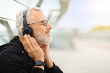Neşeli büyükbaba, yaşlı, gözlüklü, akıllı saat takan, kablosuz kulaklık takan, kapalı gözlerle müzik dinleyen, dışarıda egzersiz yaparken, yer kopyalayan yaşlı adamın yakın çekimi.