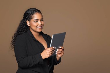 Kahverengi arka planda izole edilmiş dijital tablet kullanan neşeli Brezilyalı iş kadını, stüdyo çekimi, boş alan. İş için aygıt, çalışma, öğrenim, yatırım ve başlangıç için uygulama, reklam ve teklif