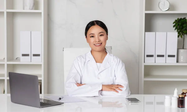 Family Doctor. Smiling Asian Therapist Lady Posing At Workplace In Office, Confident Physician Woman In Uniform Sitting With Folded Arms At Desk In Modern Clinic And Looking At Camera, Copy Space