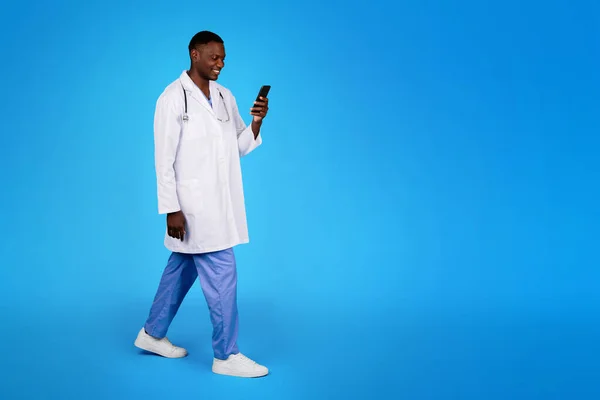 积极的年轻黑人医生穿着白衣去 用电话聊天 隔离在蓝色工作室的背景下 全身而退 医疗咨询 保健应用 广告和提供 专业咨询 — 图库照片