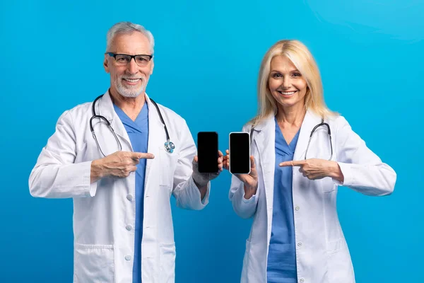 Telemedicine and Virtual Consultations. Cheerful elderly doctors man and woman show smartphones with blank screen. Remote appointments, doc diagnose patients, provide medical advice, blue background
