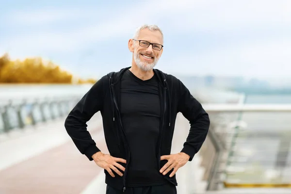Happy motivated athletic senior grey-haired sportsman wearing black sportswear jogging by street at autumn, standing on walking bridge and smiling, retired man exercising alone outdoor, copy space