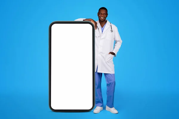 快乐的年轻黑人医生穿着白色外套靠近大智能手机 屏幕空白 隔离在蓝色的工作室背景 App Website Recommendation Health Care Medicine Remotely — 图库照片