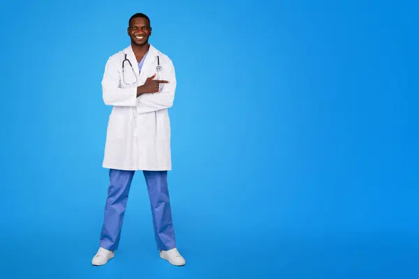很高兴有自信的年轻黑人医生身穿白衣 手指指向空旷的空间 与蓝色工作室背景隔离 关于保健 专业咨询 医疗服务的建议 — 图库照片