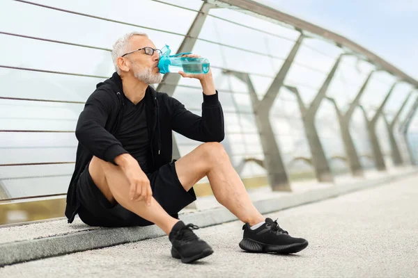 Siyah spor giyimli yaşlı yakışıklı sporcu içme suyu kullanıyor, yürüyen köprüde oturuyor, fotokopi çekiyor. Kıdemli adam dışarıda çalışır, egzersiz sırasında su içer.