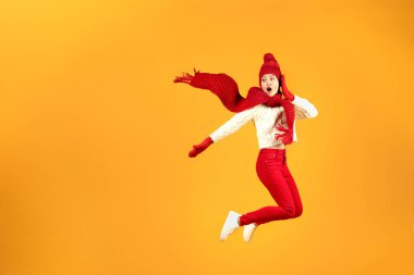Büyük Kış Teklifi. Kırmızı örgü şapkalı ve atkılı heyecanlı genç kadın sarı stüdyonun arka planına atlıyor, boş alana bakıyor, havada poz veriyor. Noel Sezonu Satış Reklamları