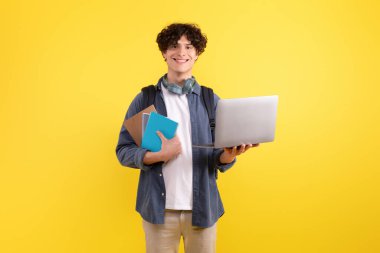 E-öğrenme. Sırt çantalı mutlu öğrenci modern dizüstü bilgisayar kullanıyor, dijital boş zamanların ve güzel eğitim sitelerinin tadını çıkarıyor, sarı stüdyo arkaplanının üzerinde duran defterlerle kameraya gülümsüyor.