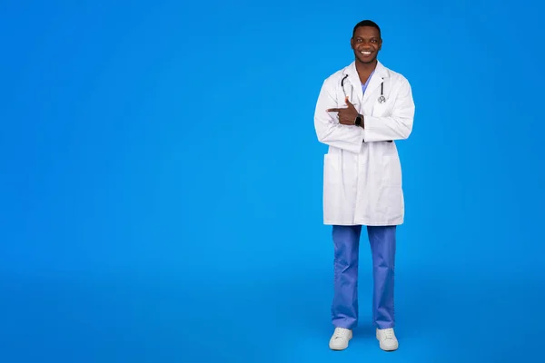 快乐的年轻非洲裔美国医生 身穿白衣 手指在空旷的空间 隔离在蓝色的工作室背景 保健建议 诊所中的药品 — 图库照片
