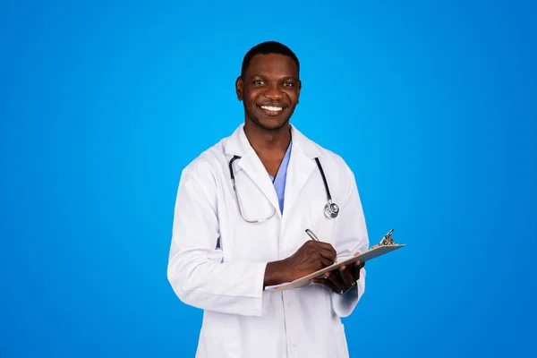 很高兴年轻的非洲裔美国医生穿着白衣 在剪贴板上做笔记 被隔离在蓝色的工作室背景下 病人人数 医疗保健 诊所治疗 — 图库照片