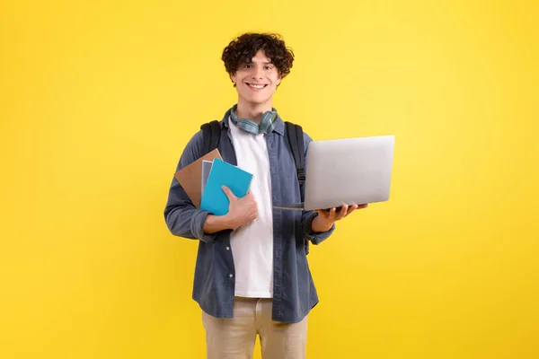 E-Learning. Happy student guy with backpack using modern laptop, enjoying digital leisure and nice educational website, standing with notebooks over yellow studio background, smiling at camera