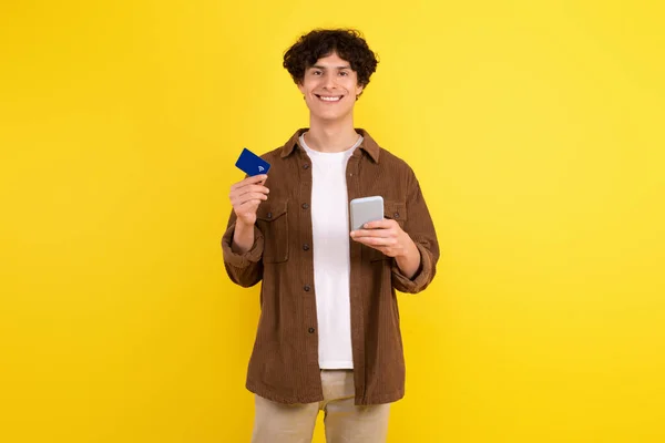 Online wallet, mobile payments. Happy guy holding credit card and smartphone in his hands, enjoying virtual shopping smiling to camera over yellow background. Ecommerce and internet banking app