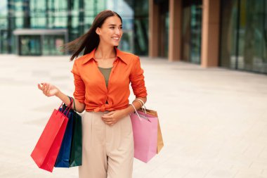 Moda yaşam tarzı. Açık havada alışveriş yapan alışveriş torbalarıyla poz veren mutlu genç bayan müşteri, şehirde yürüyüş ve alışveriş yapmaktan hoşlanıyor, mesaj atmak için boş yere bakıyor