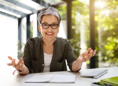 Neşeli, kısa saçlı, gözlüklü orta yaşlı bir kadın çalışanlarla çevrimiçi toplantı yapıyor, iş yerinde oturuyor, kameraya el sallıyor ve gülümsüyor, güneş parlıyor.