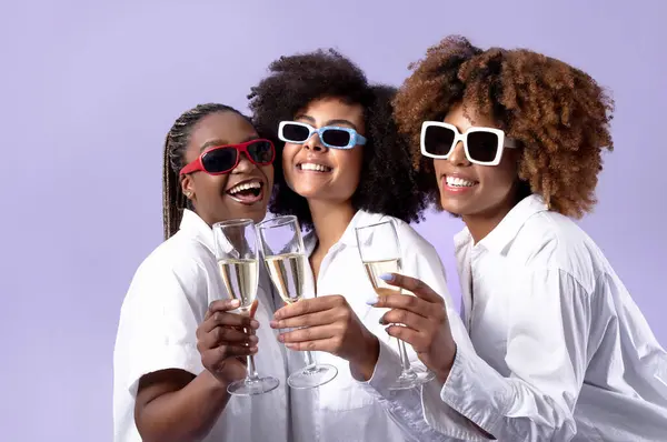 Holiday Celebration Party. Three African American Women Clinking Glasses With Sparkling Wine, Celebrating Special Event Wearing Sunglasses Standing Over Purple Studio Background