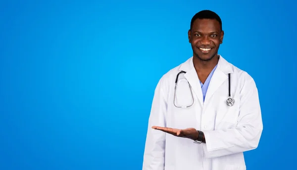 青いスタジオの背景に隔離された広告とオファーのための空のスペースを保持する白いコートの幸せな若いアフリカ系アメリカ人男性の医師 ヘルスケアサービス クリニックにおける医療の推奨事項 — ストック写真