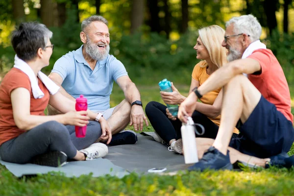 Happy senior people chatting and resting together after training outdoors, group of cheerful mature men and women relaxing outside, sitting on fitness mats in park, talking and laughing, closeup