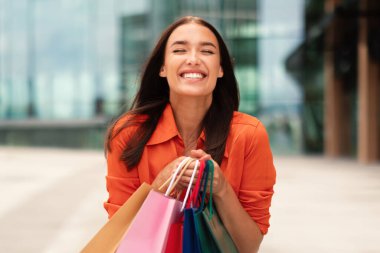 Alışveriş torbalarıyla poz veren, elinde renkli kâğıt paketler tutan, hafta sonu şehirde yürüyüş yapan ve şehir dışında ayakta duran mutlu kadın portresi. Ticaret ve indirimler