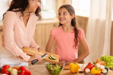 Orta Doğulu anne ve kız çocuğu birlikte sebze salatası pişiriyor, salatalıkları bowlinge ekliyor, evdeki mutfak masasında akşam yemeği hazırlıyor, kırpılmış şut çekiyor. Aile Beslenme, Sağlıklı Tarifler