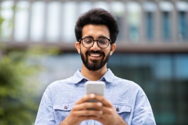 Mobil uygulama, modern teknolojiler, alet bağımlılığı. Sokakta cep telefonu kullanan, telefon ekranına bakıp gülümseyen, geçmişi bulanık, neşeli, yakışıklı, sakallı Hintli adamın yakın plan portresi.