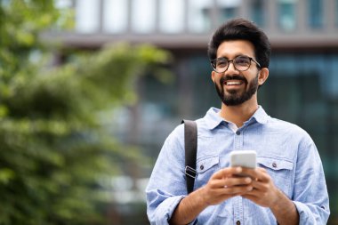 Bin yıllık mutlu Hintli adam günlük kıyafetler, gözlük takıyor, çanta taşıyor, akıllı telefon kullanıyor, fotokopi alanına bakıyor ve gülümsüyor. Güzel internet teklifi, en yeni mobil uygulama.