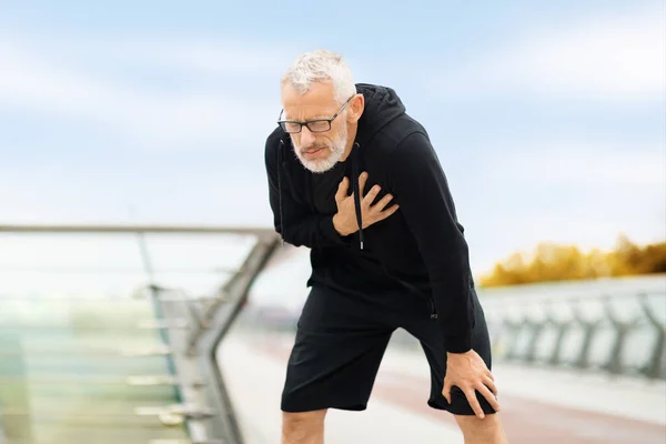 Risks for seniors during exercising. Exhausted elderly sportsman with heart pain touching his chest, jogging in the public park, copy space. Sport and cardiovascular diseases concept