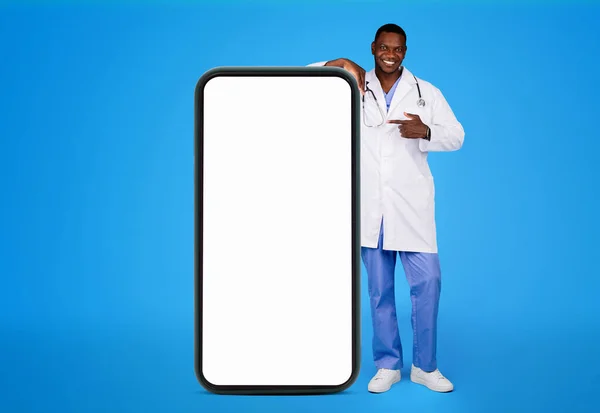 身穿白衣的年轻黑人医生对着大智能手机微笑 屏幕空空如也 隔离在蓝色的工作室背景下 远程保健推荐网站 应用程序 — 图库照片