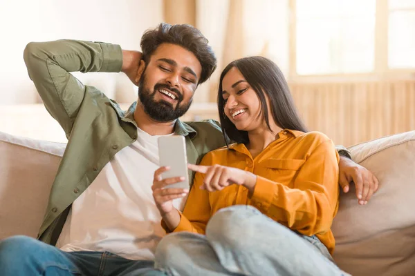 Happy young indian couple sitting on couch, using smartphone and watching funny content, lady holding gadget and pointing at screen, showing photos, sharing social media content
