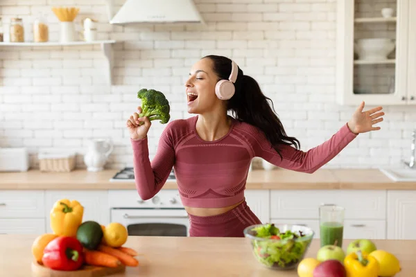 Weight Loss. Happy Fitness Lady With Headphones Singing Holding Brocoli Like Microphone, Having Fun While Cooking Healthy Slimming Recipes, Enjoying Music At Modern Kitchen. Sports Nutrition