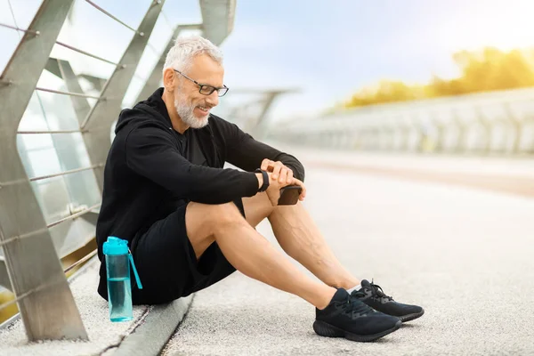 Fitness bracelet. Senior grey-haired handsome sportsman sit on the ground on walking bridge, check his smart watch, drink water while have outdoor workout, tracking his training results, copy space