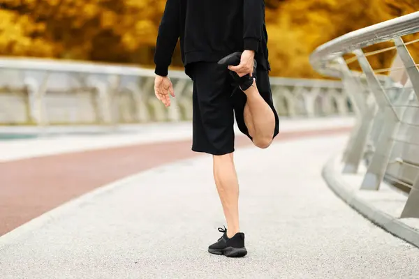 Cropped of athletic man wearing black sportswear stretching legs before jogging by walking bridge at autumn city, back view of sportsman have outdoor workout, running by park, copy space