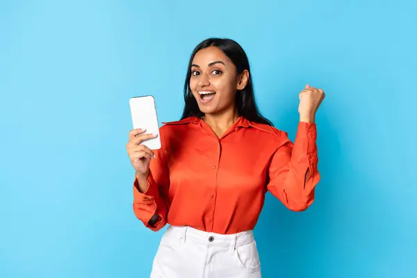 移动成功庆祝 快乐的印度年轻女性 手持智能手机 站在蓝色工作室的背景上 微笑着 站在网上庆祝胜利 — 图库照片