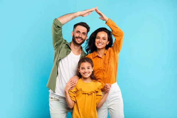 Family insurance concept. Happy european parents making symbolic roof of hands above little daughter, mother and father standing with child over blue background in studio