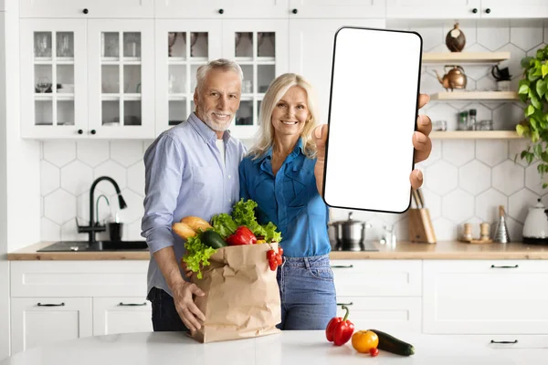 Delivery Service. Happy Mature Spouses With Grocery Bag Showing Big Blank Smartphone With White Screen At Camera, Smiling Senior Man And Woman Recommending Mobile App Or Website, Mockup