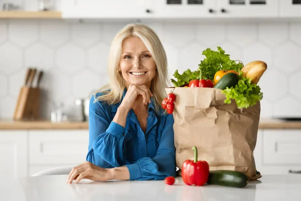 Happy Senior Woman In Kitchen Posing With Grocery Bag After Food Shopping, Smiling Mature Female Sitting At Table Next To Package With Fresh Vegetables, Enjoying Healthy Eating, Copy Space