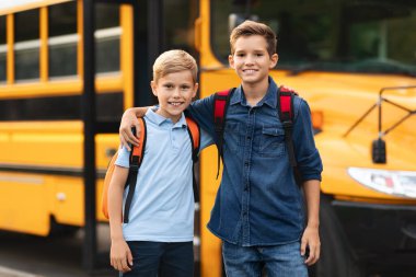 Dışarıda okul servisinin yanında iki mutlu çocuğun portresi, dışarıda duran sırt çantalı neşeli çocuklu çocuklar, sınıf arkadaşları kameraya sarılıp gülümsüyor, birlikte çalışmanın keyfini çıkarıyorlar.