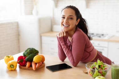 Sports Nutrition Application. Happy Sporty Lady Standing Near Table With Digital Tablet, Fruits And Vegetables, Using App For Slimming And Weight Loss Posing In Fitwear At Modern Kitchen At Home clipart