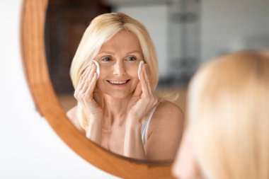 Çekici olgun bir kadının yakın plan portresi aynanın önünde pamuklu bez ile cildini temizliyor orta yaşlı bir kadın evde kendine bakmaktan zevk alıyor, yansımasına gülümsüyor.