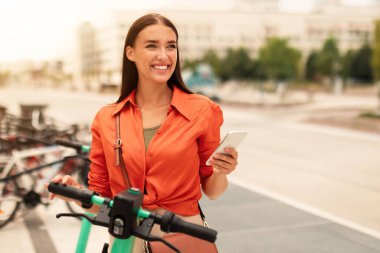 Neşeli genç bayan, elektrikli scooter 'ın kilidini açıyor, cep telefonu uygulaması kullanıyor, modern şehirde akıllı toplu taşıma için Eco taşıma aracı kiralıyor, dışarıdaki sokakta duran bisiklet parkında duruyor.