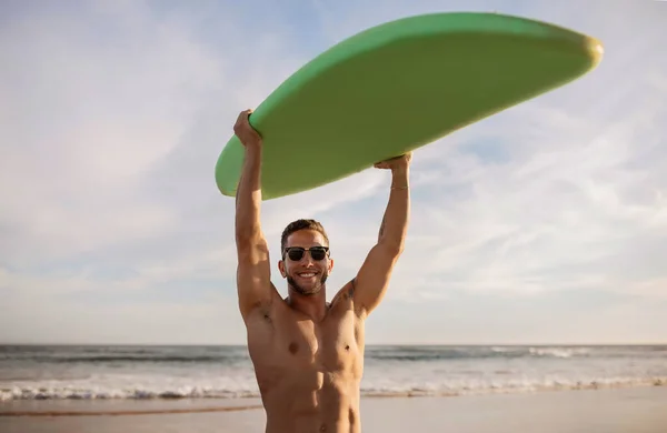 ビーチにポーズしながら頭の上にサーフボードを運ぶ運動的な若いシャツレスサーファーマー カメラを見てスタイリッシュなサングラスでハンサムなミレニアル男 サーフィンや夏休みを楽しむ — ストック写真