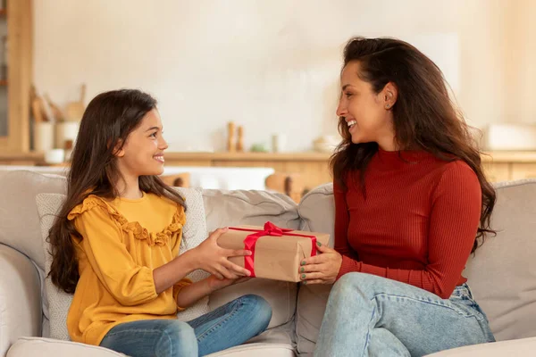 Surprise Gift For Daughter On Birthday. Cheerful Arab Mom Giving Wrapped Present Box To Preteen Daughter, Sitting On Sofa, Congratulating And Celebrating Family Holiday At Home