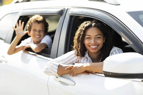 Happy black mother and child looking from white car windows, waving and smiling at camera. Cheerful african american family enjoying road trip by brand new auto, going vacation, weekend journey